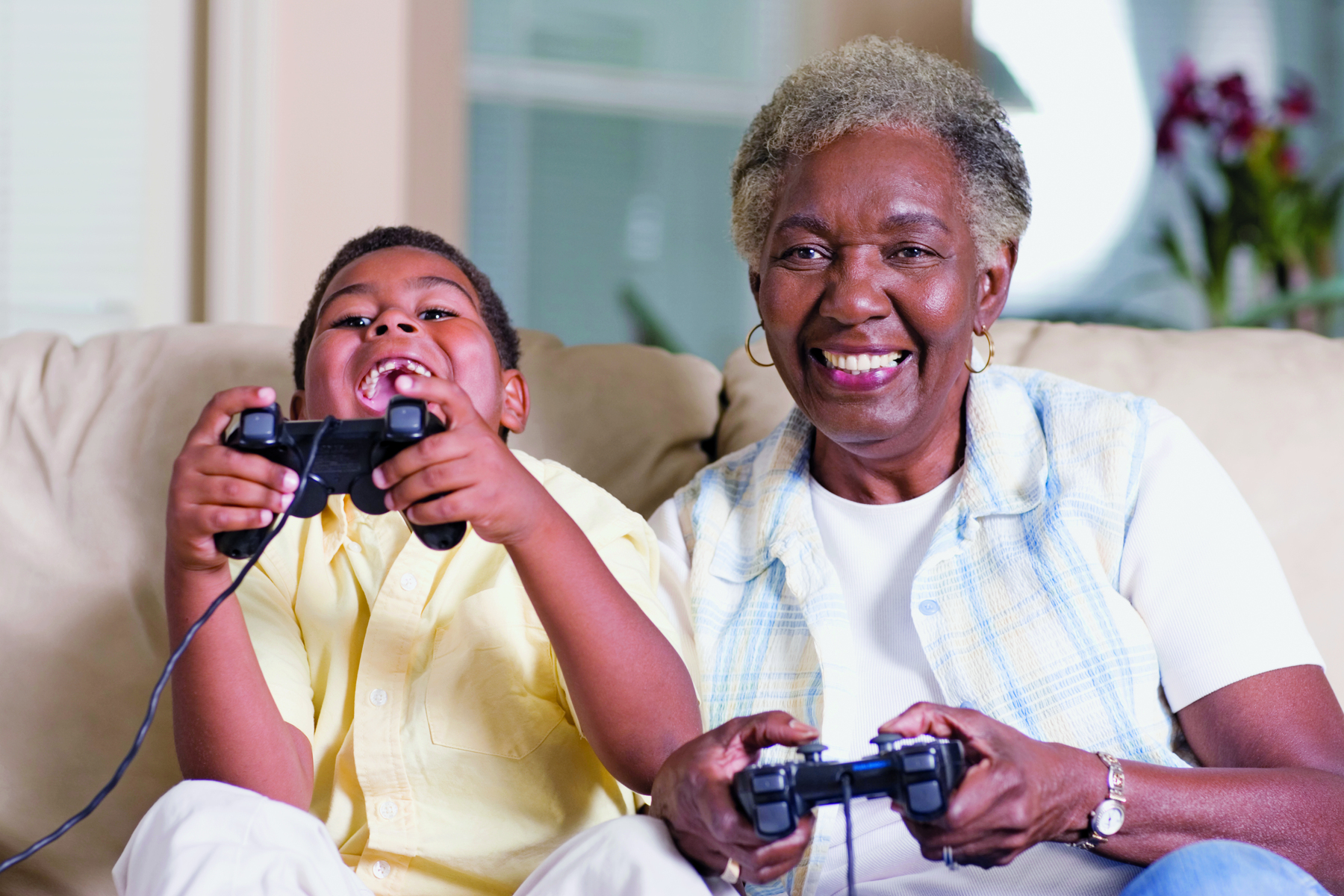 Fotografia. Uma criança e uma mulher idosa, de cabelo grisalho, estão sentadas no sofá, jogando videogame e sorrindo. Cada uma tem um console na mão.