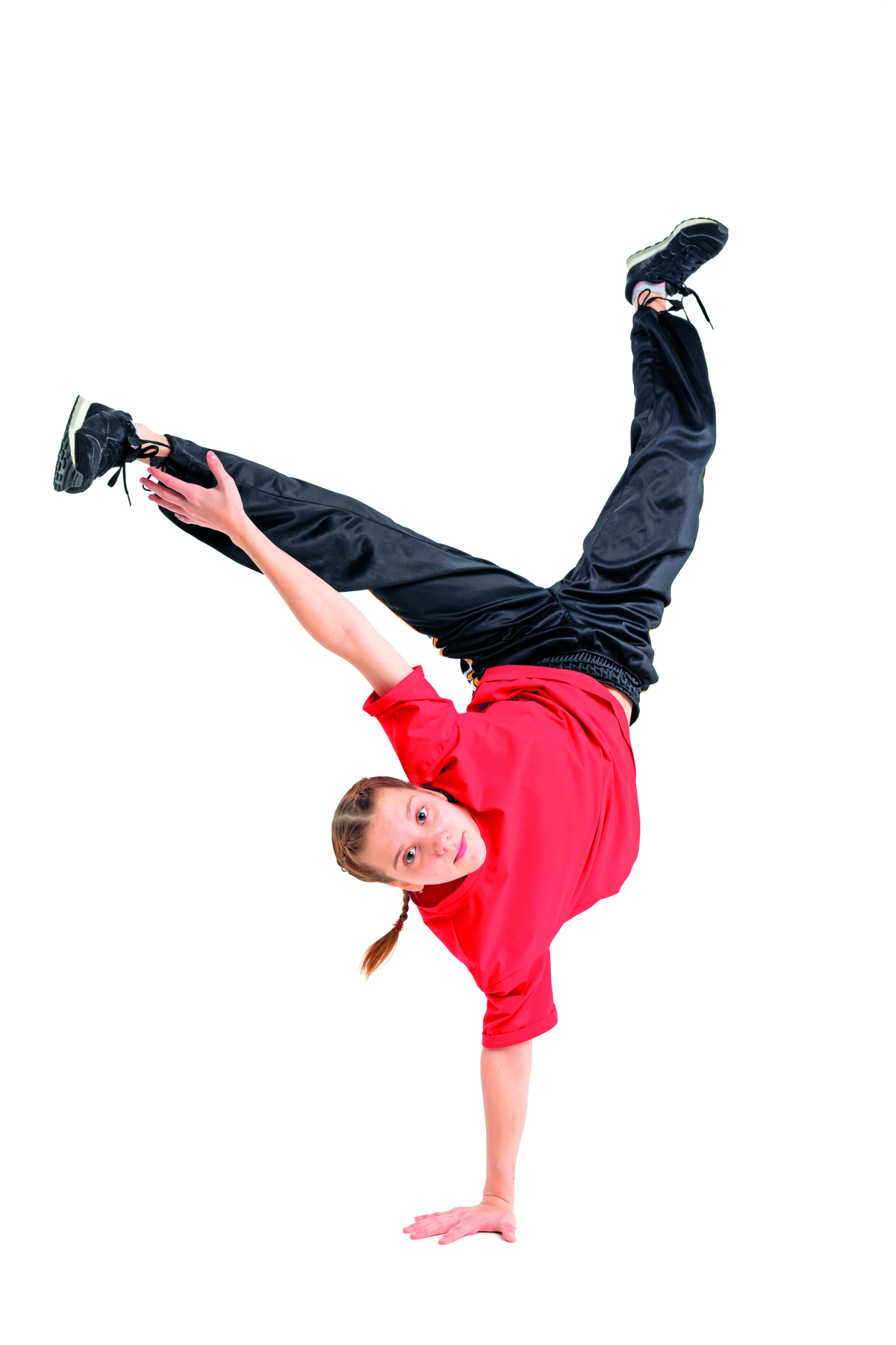 Fotografia. Menina usando blusa vermelha, calça e tênis pretos. Ela está com a mão direita apoiada no chão, as duas pernas levantadas e afastadas e com o braço esquerdo esticado na direção do pé.
