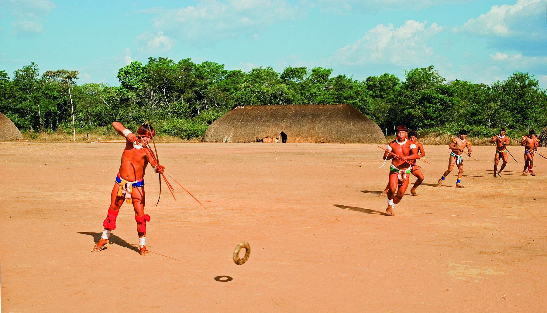 Fotografia. Homem indígena está em pé e segura um arco e flecha apontado na direção de um objeto circular que paira acima do chão de terra. Ao fundo, há uma oca, vegetação e diversos indígenas.