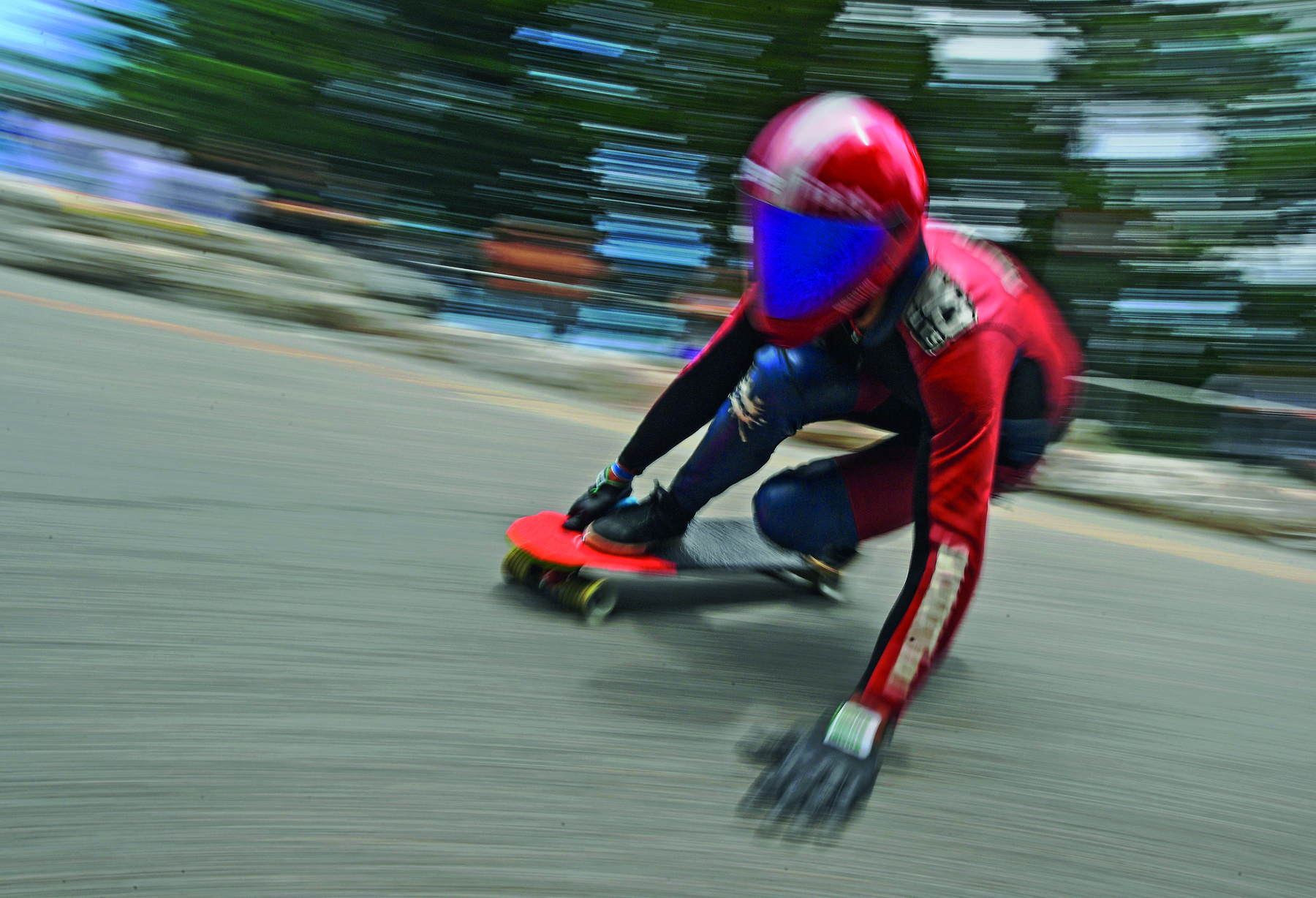 Fotografia. Pessoa usando capacete vermelho espelhado, camiseta vermelha e tênis pretos sobre um skate. Ela está agachada, com a mão direita no skate e a mão esquerda ao lado. A fotografia nos permite concluir que ela desce por um terreno em grande velocidade.
