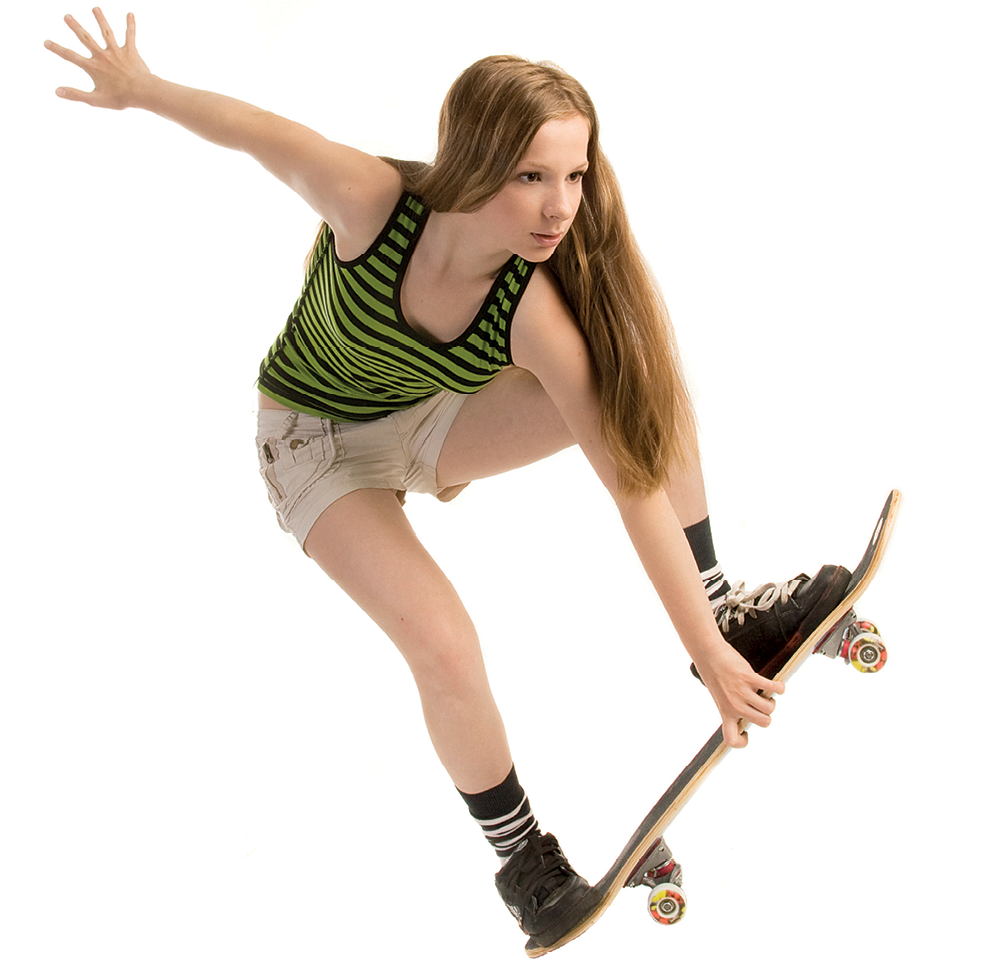 Fotografia. Garota, de regata verde e  preta listrada e short bege, se equilibra em cima de um skate. O braço esquerdo segura o skate e o direito está voltado para trás, com a palma da mão aberta.