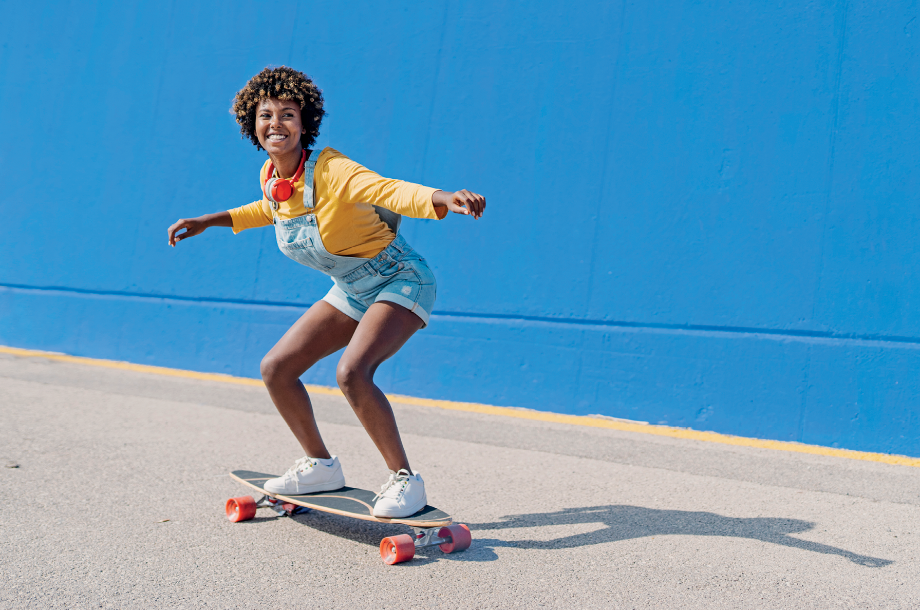Fotografia. Mulher usando camiseta amarela e macacão jeans em pé sobre um skate com os joelhos dobrados e os braços abertos.