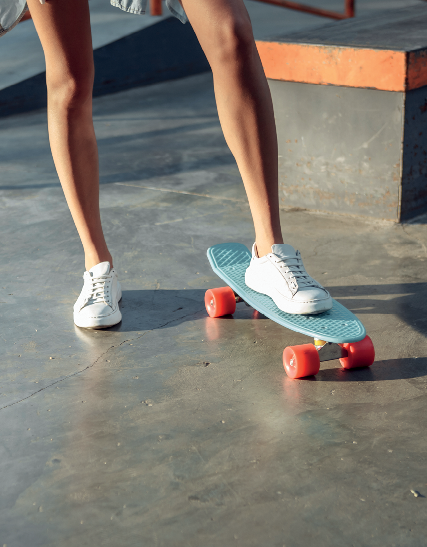 Fotografia. Destaque para duas pernas usando tênis branco e com um pé sobre um skate pequeno, azul e com rodas grossas e vermelhas.