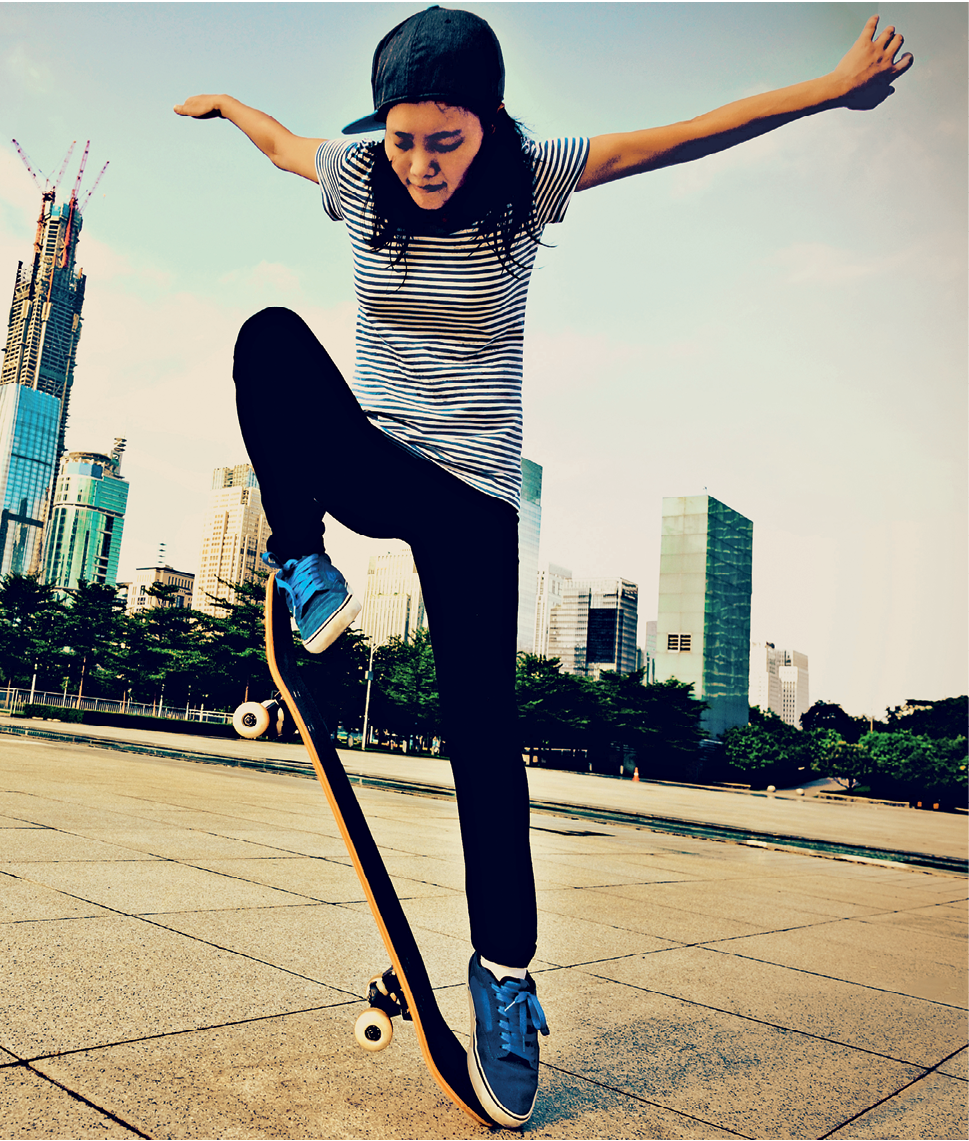 Fotografia. Mulher de boné preto, camiseta listrada, calça preta e tênis azuis saltando sobre um skate com a perna direita dobrada e o pé inclinado. A perna esquerda está esticada e os dois braços, abertos.