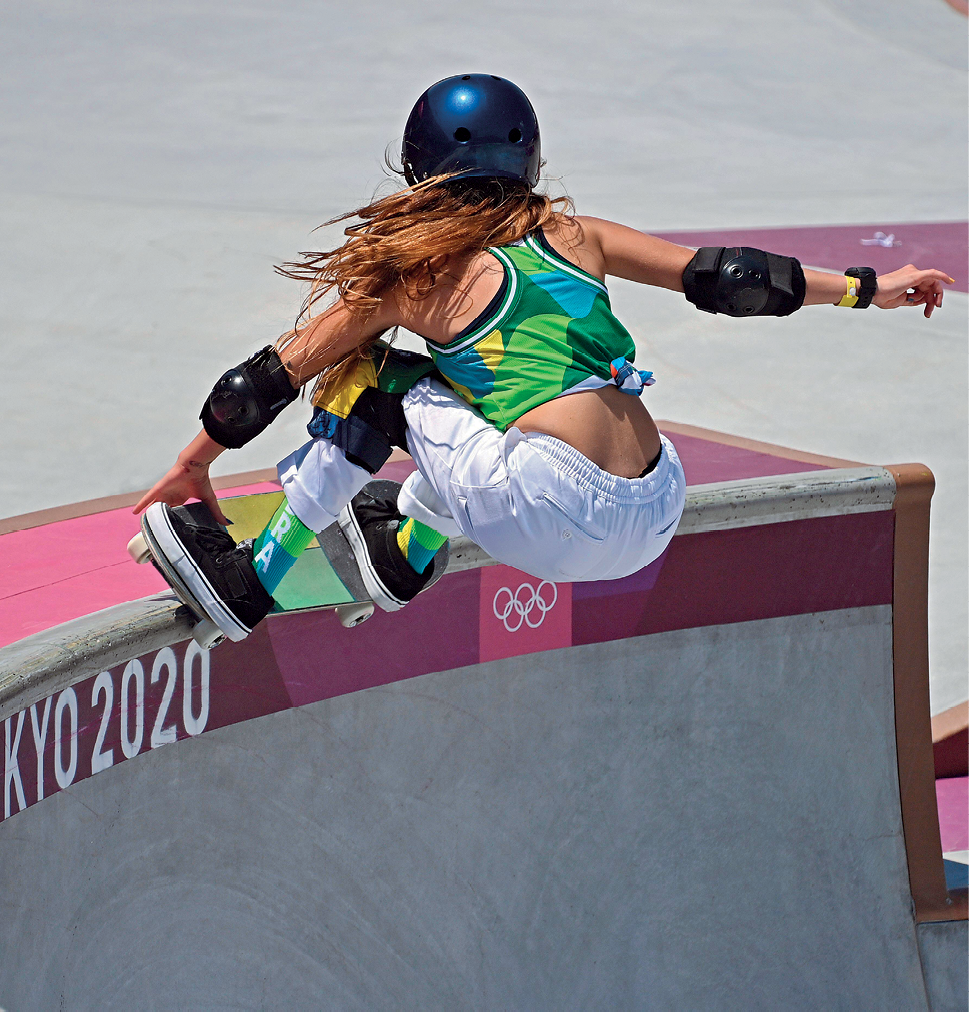 Fotografia. Mulher usando capacete azul-escuro, regata verde, calça branca e tênis pretos está com as pernas dobradas para frente sobre um skate no alto de uma rampa. Sua mão esquerda está no shape do skate.
