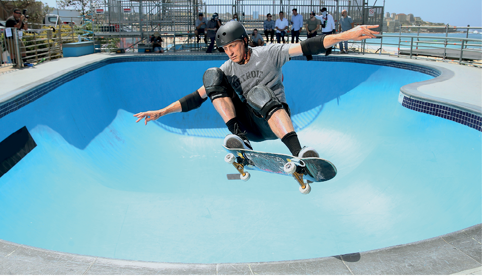 Fotografia. Um homem usando capacete preto, cotoveleiras, joelheiras, camiseta cinza, está saltando sobre um skate no alto de uma rampa e com os braços abertos; ao fundo há uma pista com formato de piscina com as bordas altas.