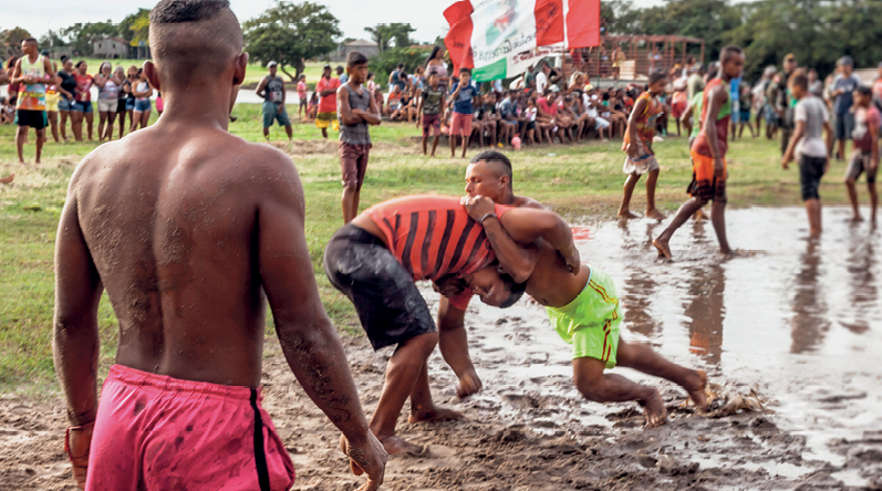 Fotografia. Dois homens lutam em um terreno com lama. Um está inclinado sobre o outro, tocando-se com os braços. As pernas de um estão flexionadas, a do outro, esticadas. Há várias pessoas ao redor, observando.