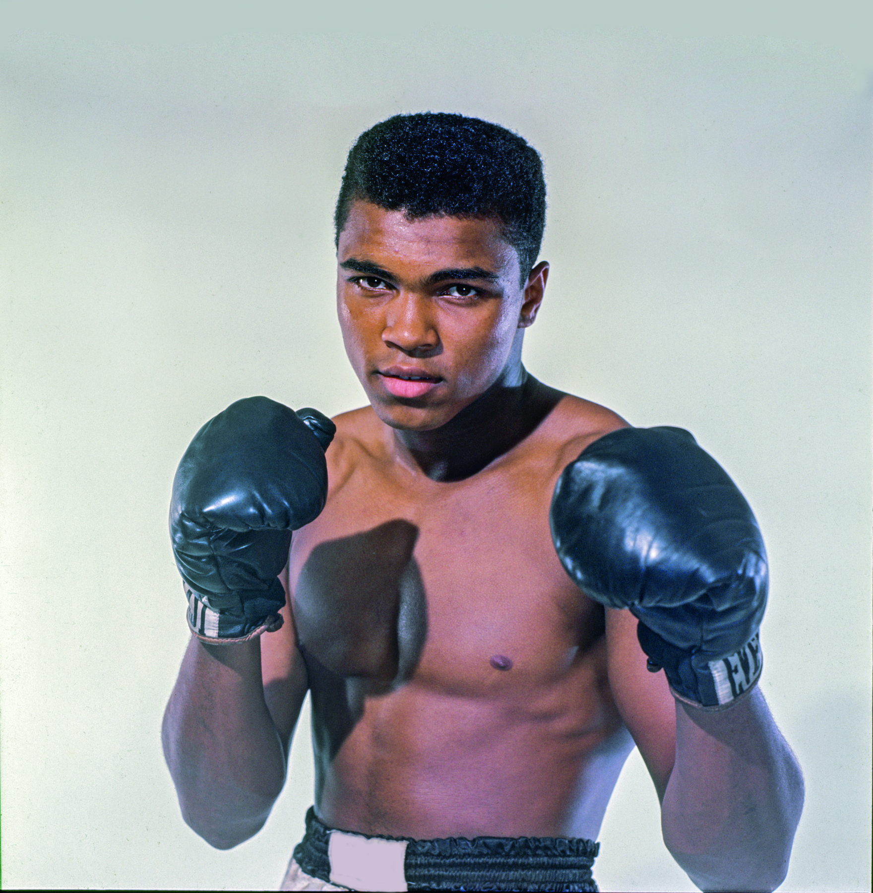 Fotografia. Boxeador Cassius Clay, usando luvas e calção pretos com os dois braços dobrados à frente do corpo.