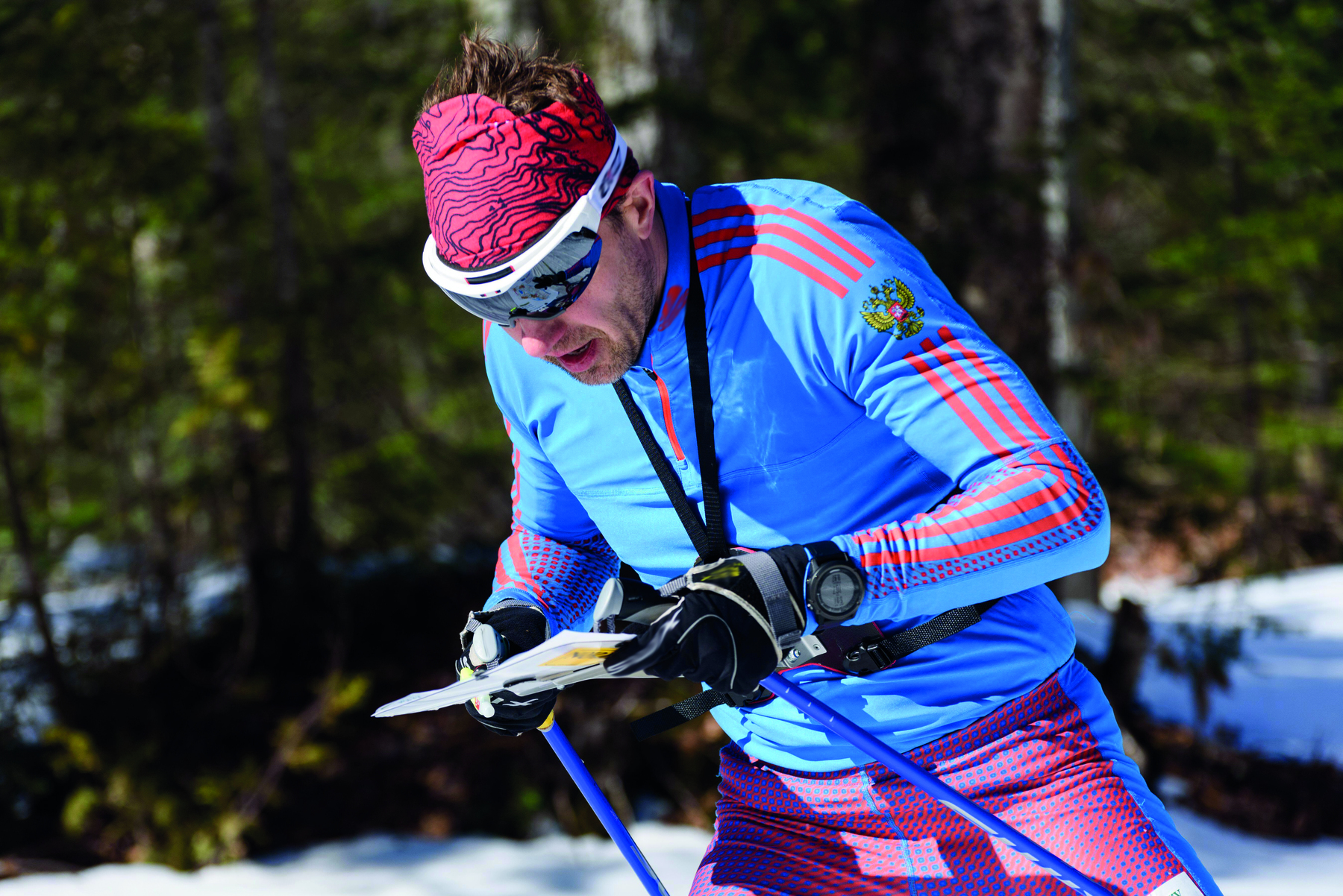 Fotografia. Homem usando óculos escuros, blusa azul, luvas pretas e bastões de esqui observa um mapa, que segura com as duas mãos.