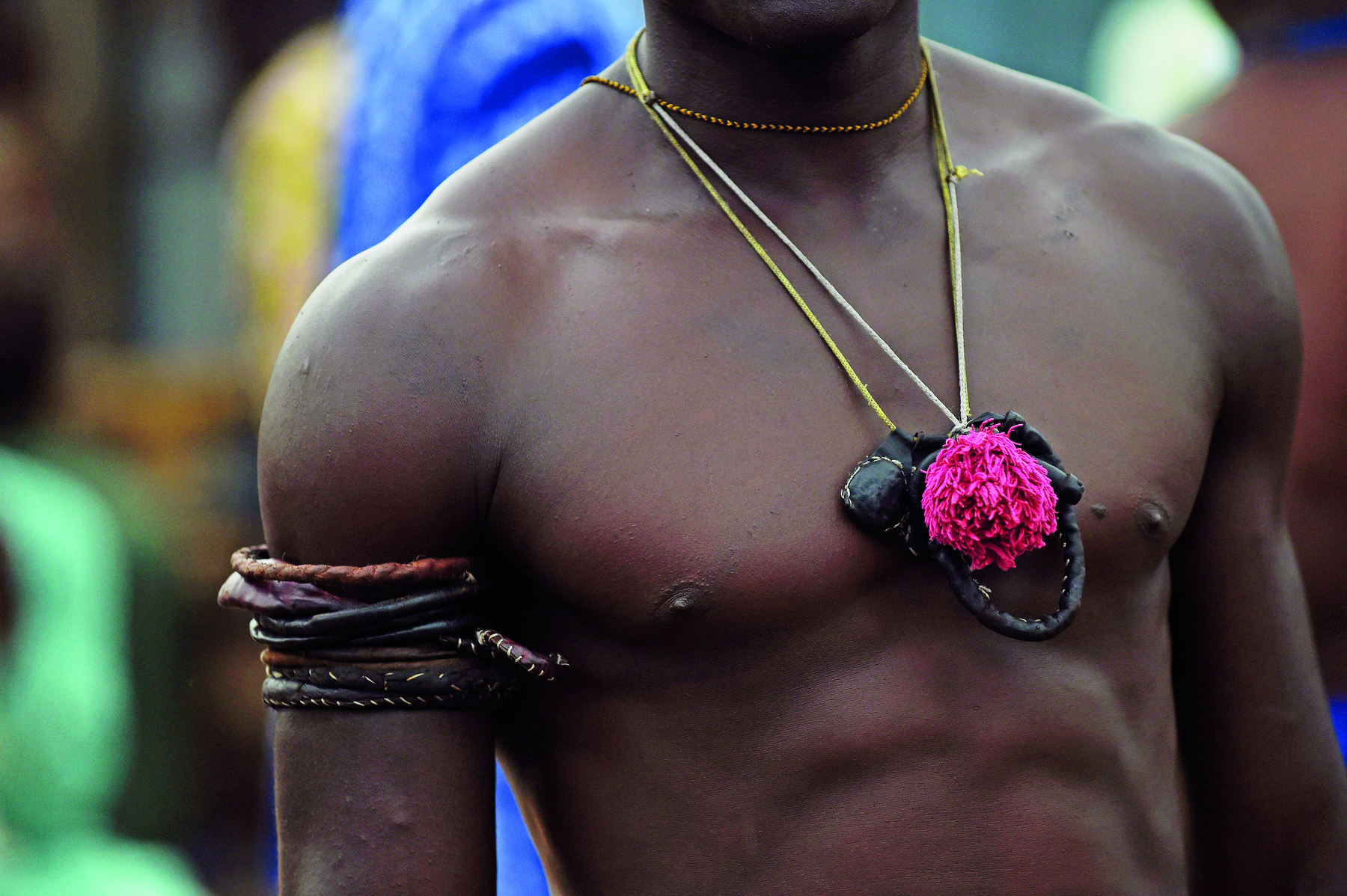 Fotografia. Homem sem camiseta usando um amuleto preto e vermelho no pescoço. Em seu braço direito há adornos com cordões coloridos.
