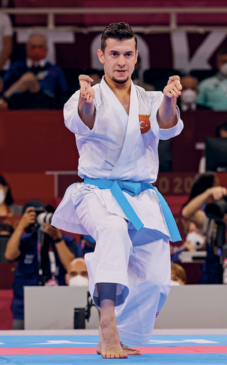 Fotografia. Homem usando quimono branco com faixa azul na cintura com a perna direita na frente e os dois braços esticados para frente com o punho fechado.
