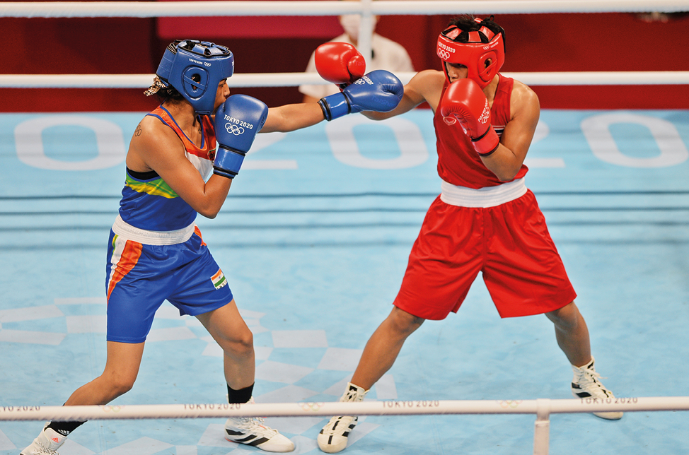 Fotografia. Duas mulheres lutando boxe. Uma está usando capacete, uniforme e luvas azuis; outra, usando capacete, uniforme e luvas vermelhas. As duas estão em pé, com os joelhos levemente flexionados, um dos braços dobrado ao lado do rosto e o outro esticado na direção do rosto da adversária.