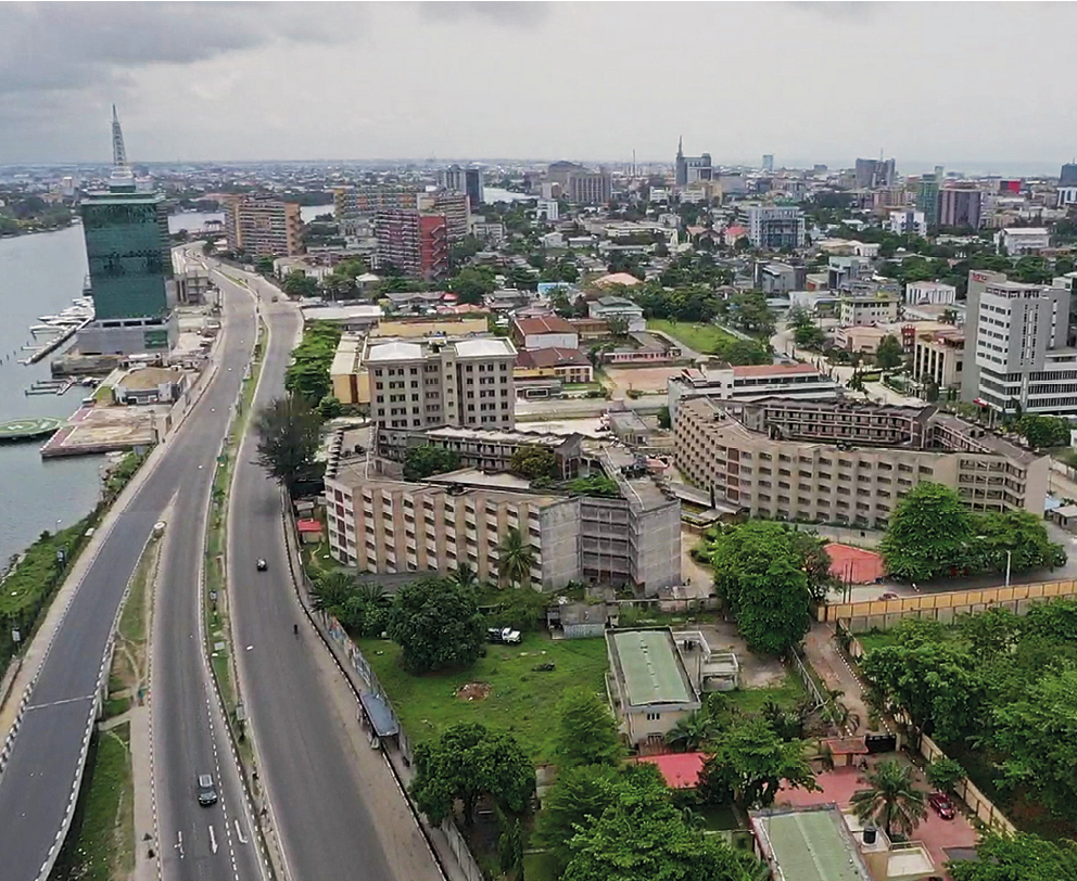Fotografia. Vista aérea da cidade de Lagos. À esquerda, um rio e uma estrada. No centro e à direita, prédios e praças com árvores.