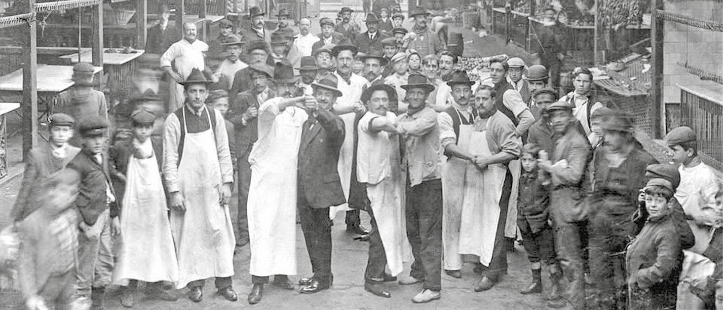 Fotografia em preto e branco. À frente, em um mercado antigo, homens de chapéu, roupa social e avental estão de mãos dadas, dançando. Ao redor, há outros homens e crianças.