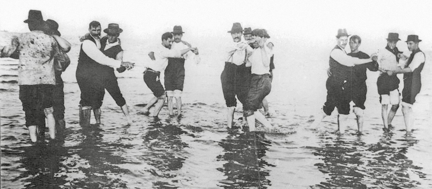 Fotografia em preto e branco. Seis duplas de homens usando roupa social com coletes ou suspensórios e chapéus. Eles estão de mãos dadas, dançando na parte rasa de um rio, com as calças arregaçadas.
