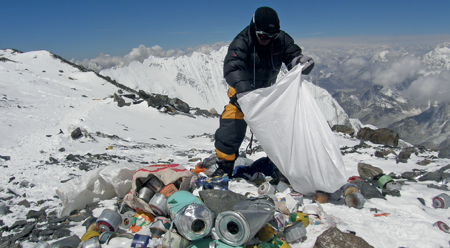 Fotografia. Homem de touca, jaqueta e calça preta segurando um saco de lixo aberto. Ao redor dele, há diversas embalagens descartadas e entulhos. Ao fundo, montanhas cobertas por neve.