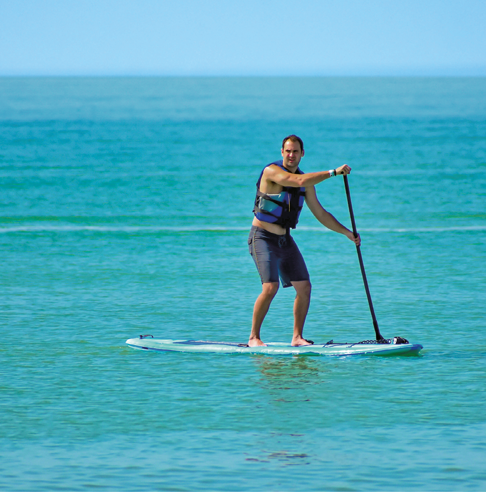 Fotografia. Um homem de cabelo preto curto usando colete salva-vidas azul e bermuda cinza, descalço, está em pé sobre uma prancha larga no mar segurando com as duas mãos um remo na direção da água.