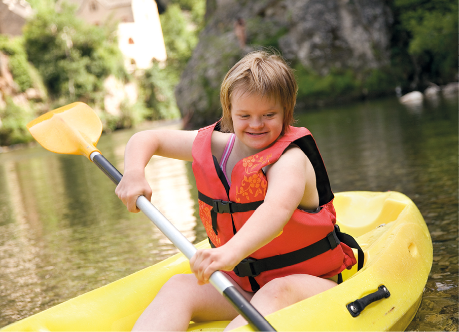 Fotografia. Uma menina de cabelos loiros curtos, olhos oblíquos e rosto redondo usando colete salva-vidas laranja está sentada em uma canoa amarela na água e segura um remo. Ao fundo, vê-se a margem, com rocha e vegetação.