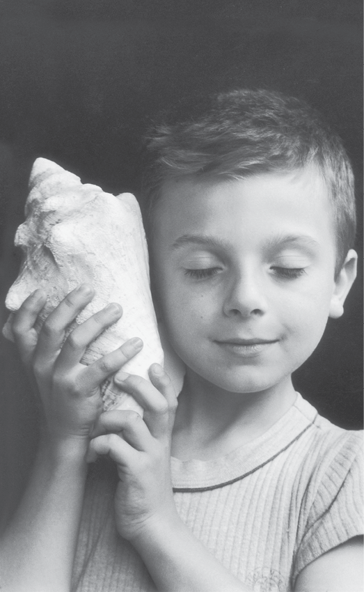 Fotografia em preto e branco, destacando o busto de um menino com cabelos curtos. Ele está com os olhos fechados e com as mãos segura uma concha encostada ao  ouvido direito.