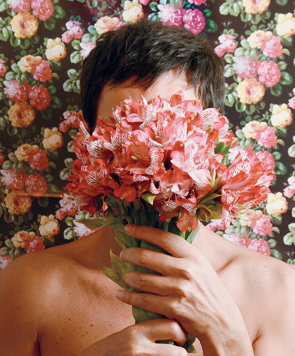 Fotografia. Um homem com cabelos curtos e escuros segura um buquê de flores de cor rosa, cobrindo o rosto. Ao fundo, diversas flores nas cores rosa e amarela.