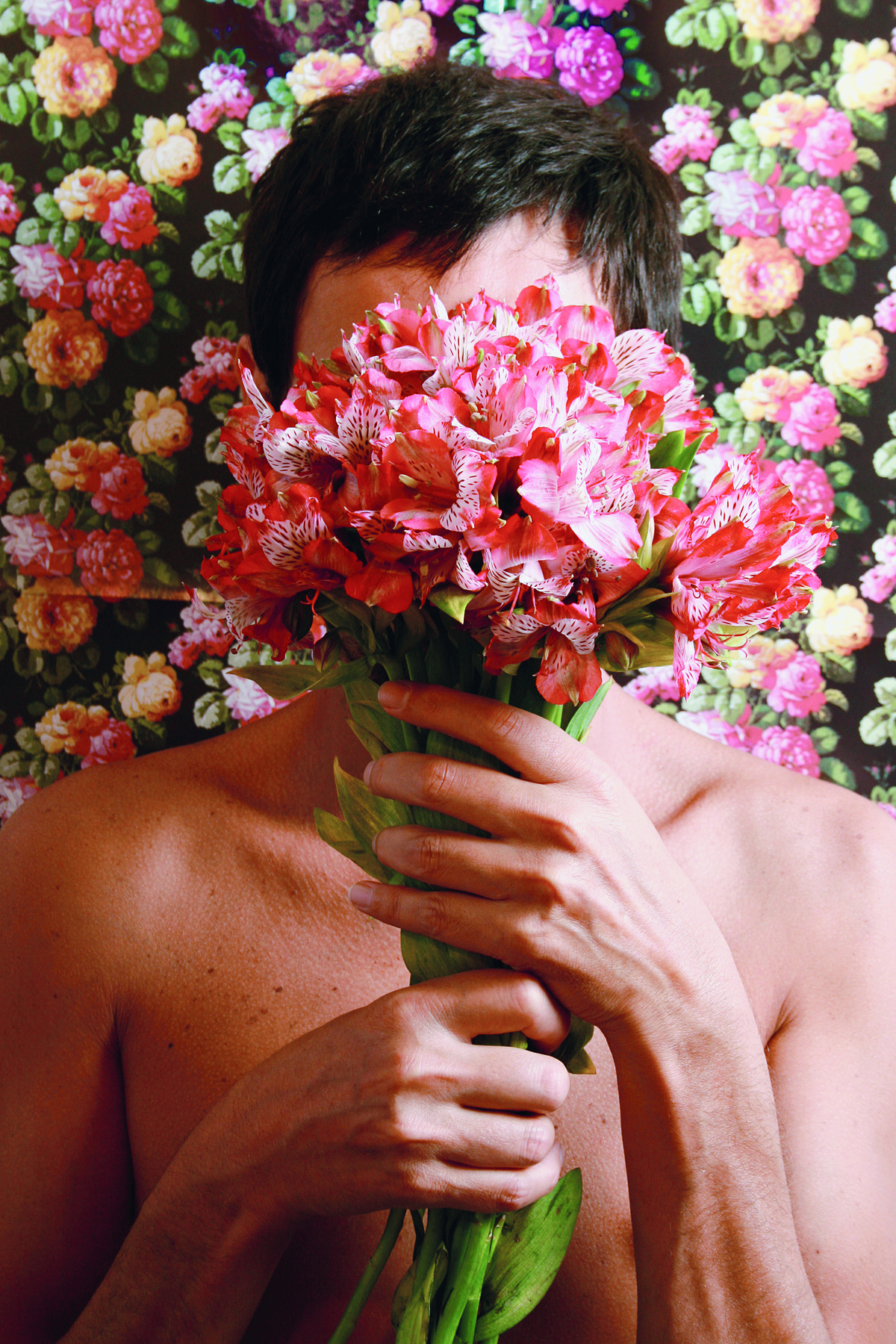 Fotografia. Busto de um homem com cabelos curtos, segurando com as mãos um buquê de flores de cor-de-rosa, cobrindo o rosto. Atrás dele, um mural com diversas flores em cores rosa e amarela.