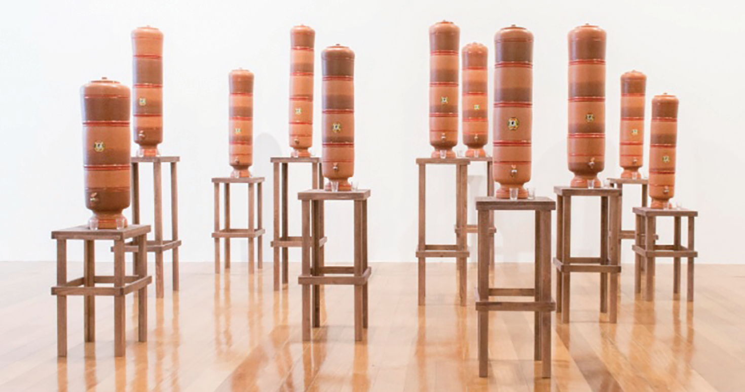 Fotografia. Uma instalação artística. Sobre um piso de madeira, onze filtros  filtros de cerâmica sobre bancos de madeira de alturas variadas.