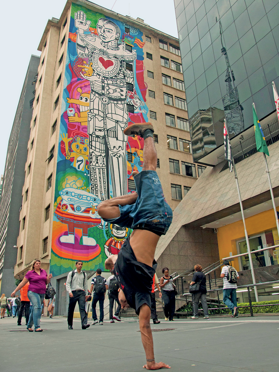 Fotografia. Um jovem fazendo um passo de dança na rua,  de cabeça para baixo, apoiado no braço direito e com a perna direita estendida para cima. Ao fundo, pessoas andando na rua, edifícios, um deles com um  grafite na fachada, composto por desenho de um homem em preto e branco com um coração vermelho. Em torno dele,  diversas criaturas e formas coloridas.