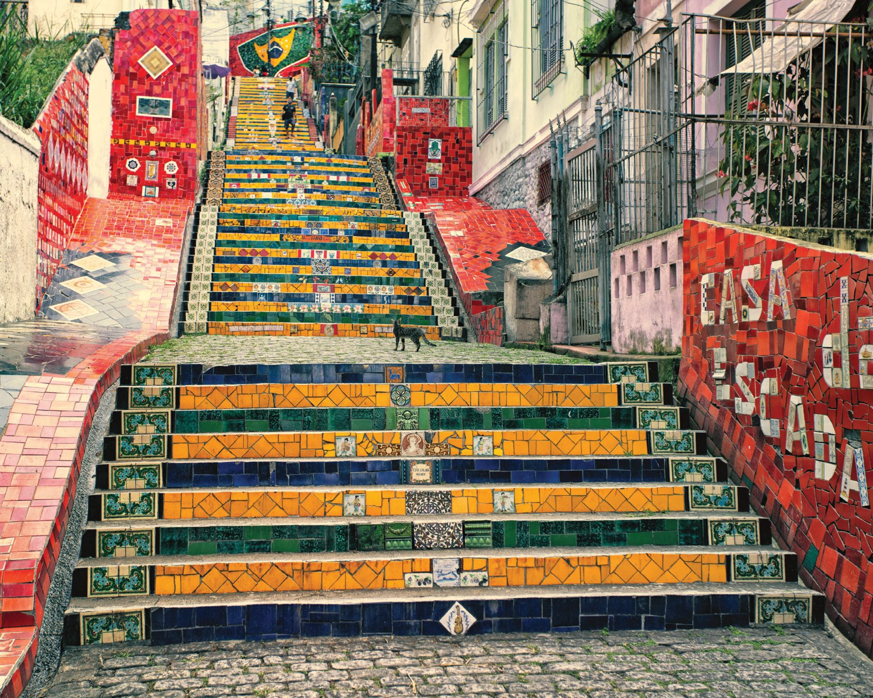 Fotografia. Destaque de uma escadaria com mosaicos amarelos, verdes e azuis nos espelhos dos degraus. Nas paredes, mosaicos  vermelhos com detalhes coloridos.