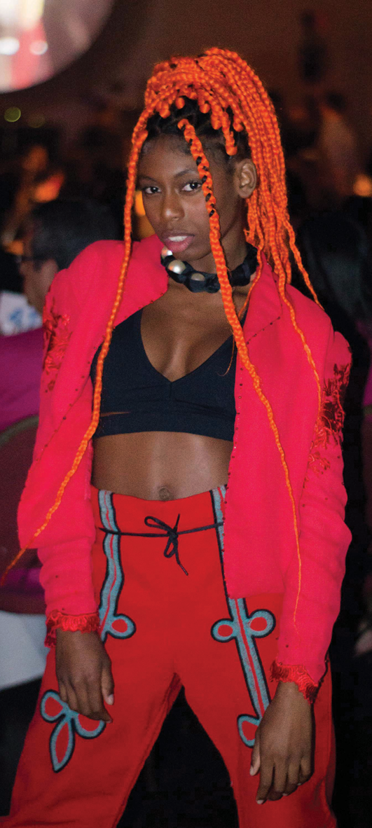 Fotografia. Mulher jovem negra com cabelos com dreads vermelhos, presos no alto da cabeça. Ela veste um top preto, casaco e calça vermelhos.