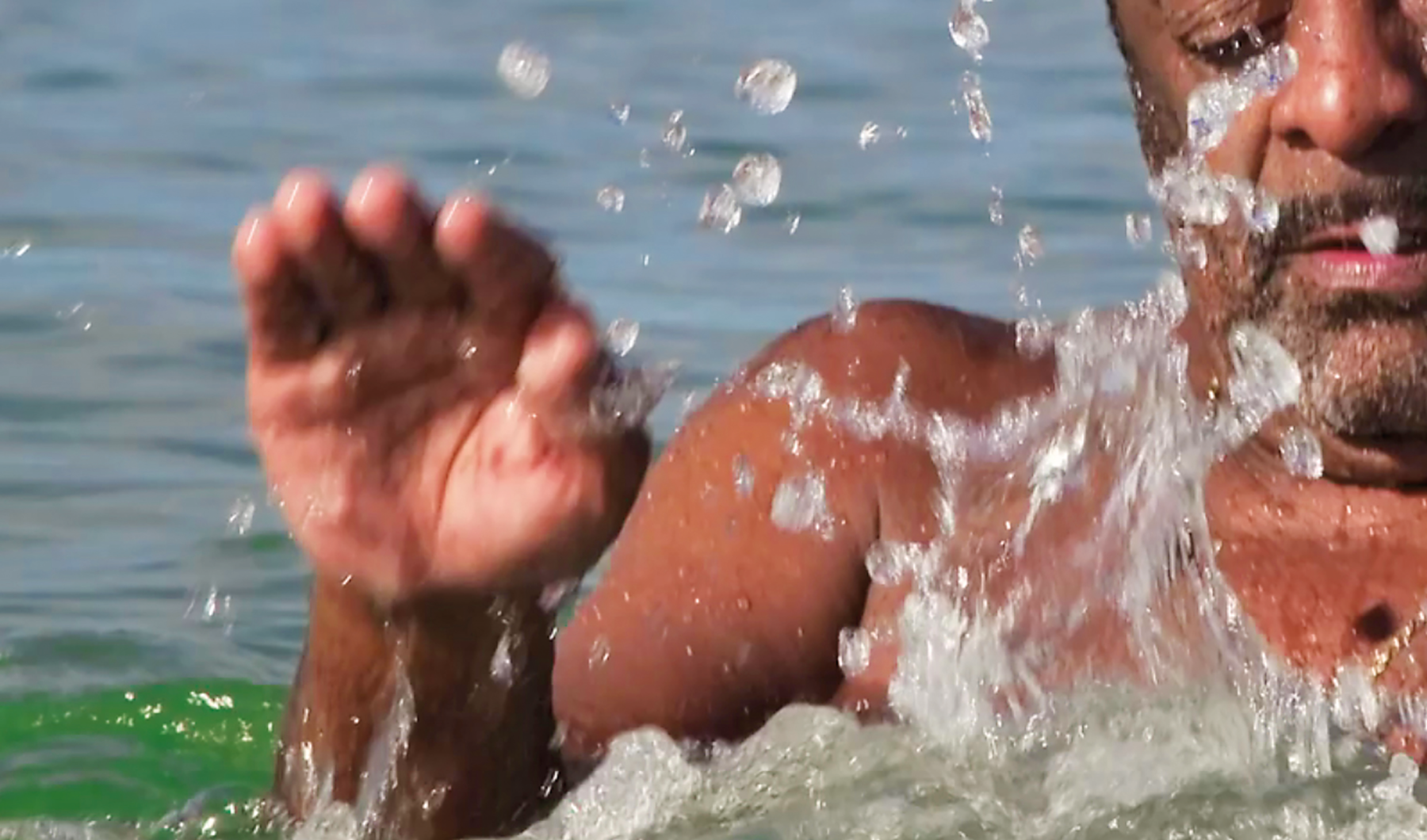 Fotografia. Detalhe do busto de um homem negro, de barba,  dentro do mar. Ele está com a mão direita espalmada na direção da superfície da água, que espira formando gotículas.
