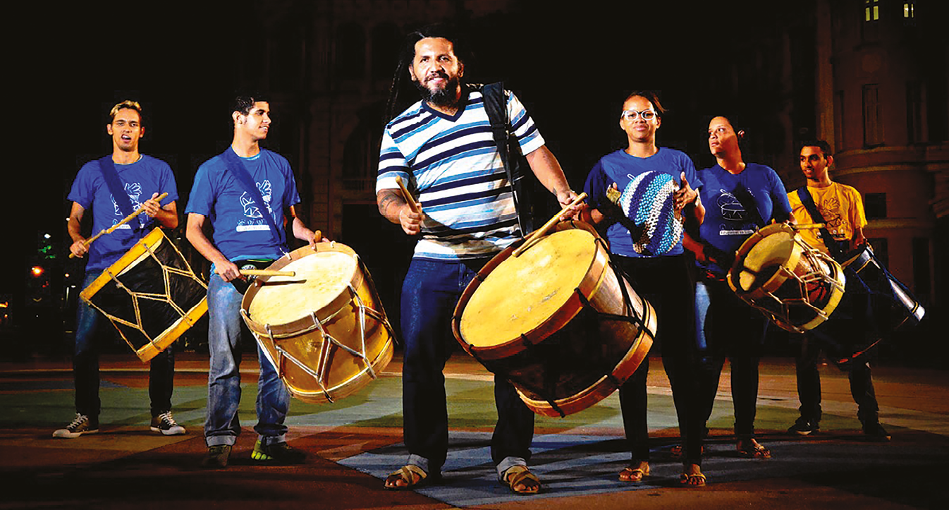 Fotografia. Seis músicos em  um palco, homens e mulheres, vestindo jeans e camisetas azuis e amarelas. Eles seguram tambores e baquetas.
