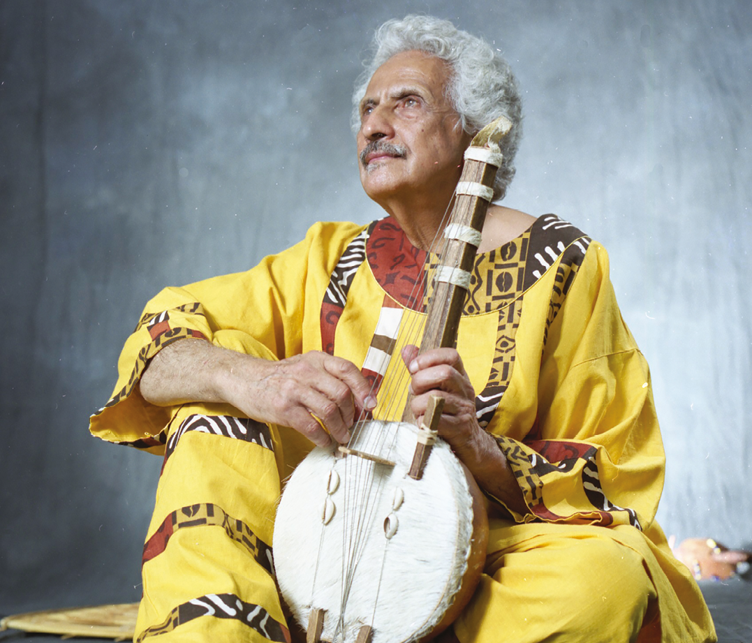 Fotografia. Um homem com cabelos brancos e bigode, usando uma túnica e calça amarelas com estampas africanas. Ele está sentado segurando um instrumento de corda.
