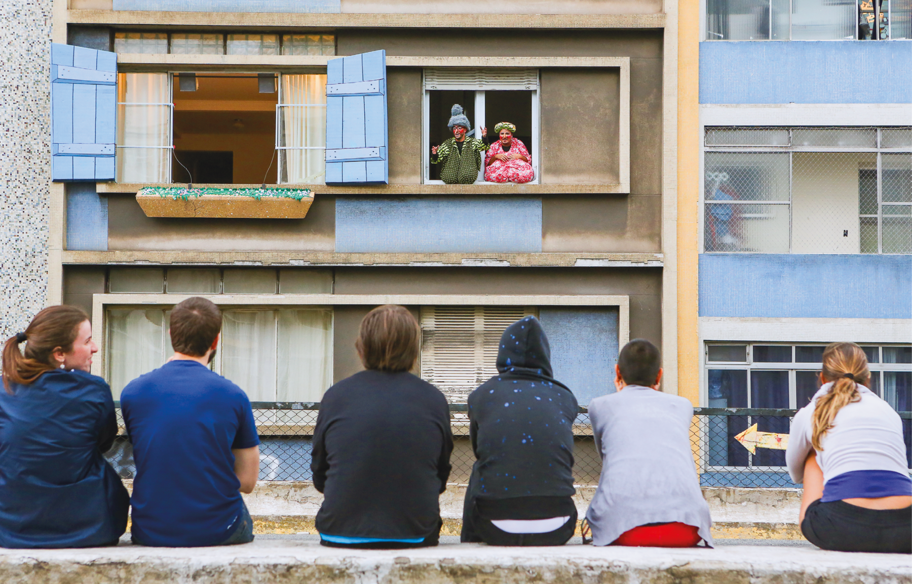 Fotografia. Em primeiro plano, pessoas sentadas em uma mureta, de costas, assistem a um espetáculo. Ao fundo, prédios com janelas. Em uma das janelas abertas, um homem e uma mulher com fantasias se comunicam com os espectadores.