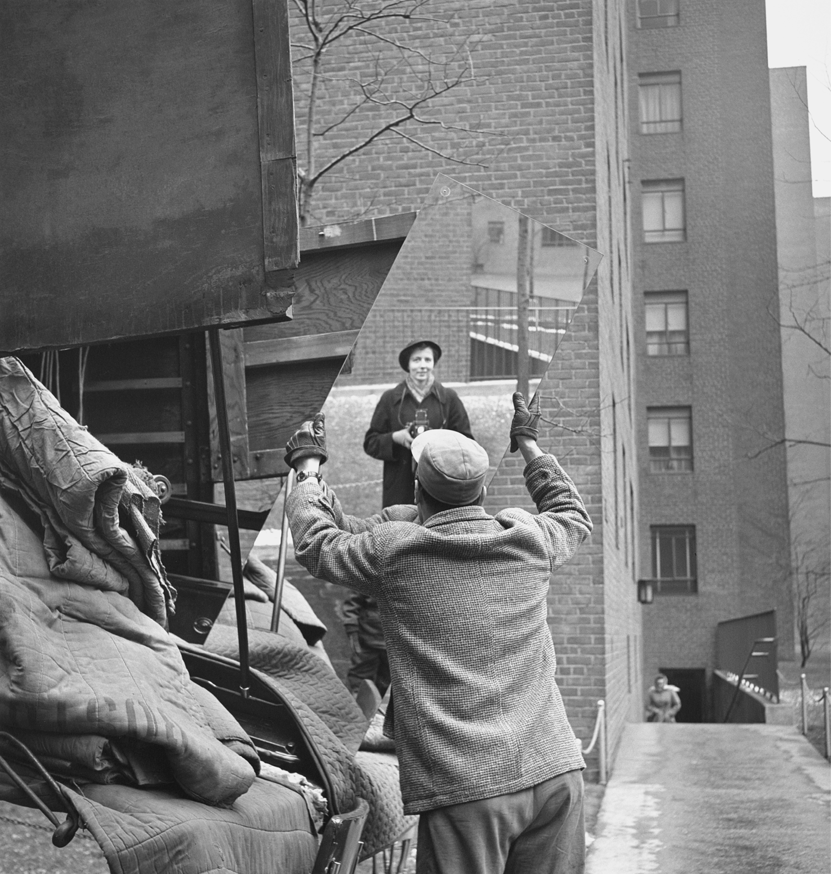 Fotografia em preto e branco. Um homem usando, casaco, boné e luvas. Ele está segurando um espelho com as mãos. Refletida no espelho, imagem de uma mulher usando chapéu e casaco, segurando com as mãos uma máquina fotográfica antiga.