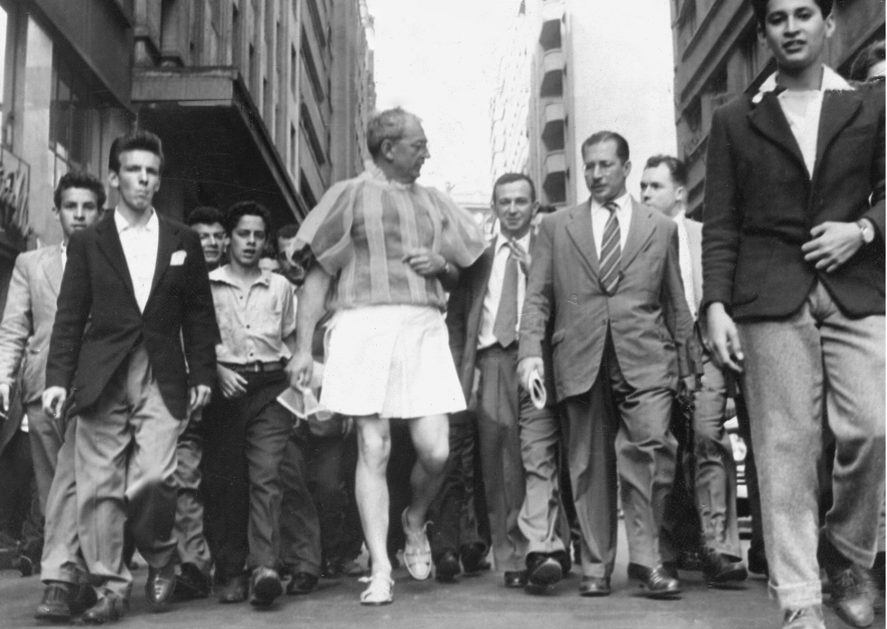 Fotografia em preto e branco. No centro da imagem, um homem grisalho e de óculos usa uma blusa bufante, saia e sandálias. Ao redor, diversos homens usando terno. Eles estão caminhando em uma rua. Nas laterais, prédios.