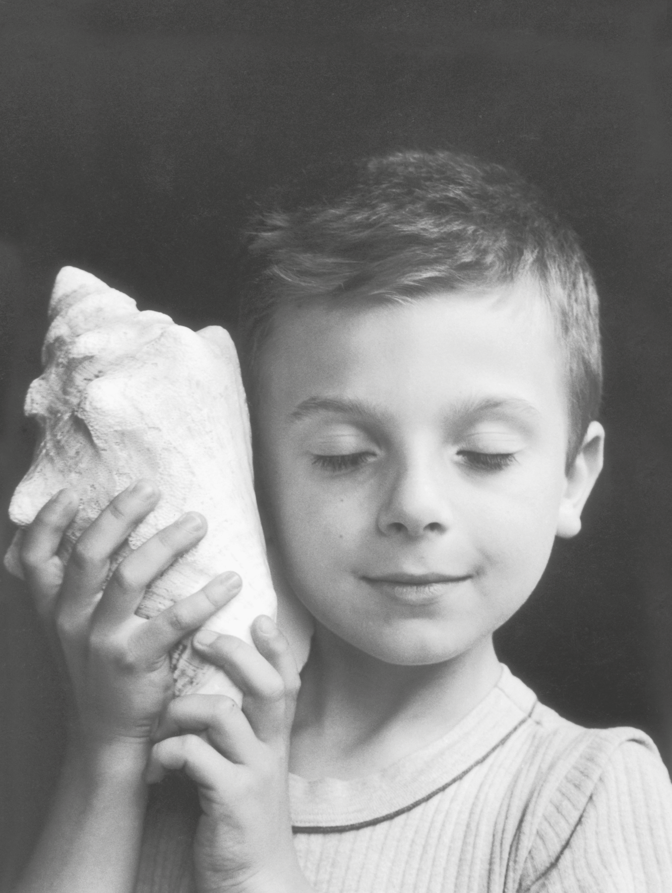 Fotografia em preto e branco, destacando o busto de um menino com cabelos curtos. Ele está com os olhos fechados e com as mãos segura uma concha encostada ao  ouvido direito.