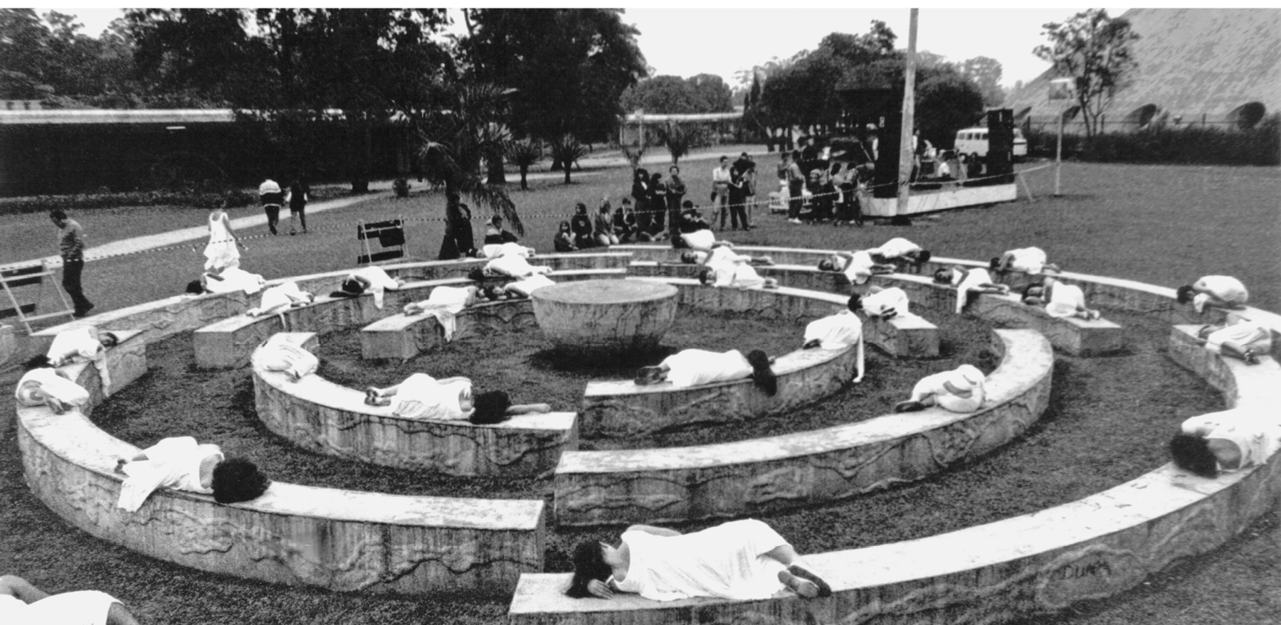 Fotografia em preto e branco. Um parque, com bancos  em círculos concêntricos e gramado. Sobre os bancos, pessoas usando roupas brancas. Algumas estão encolhidas e outras deitadas sobre os bancos.  Ao fundo, há pessoas sentadas e outras de pé assistindo à performance.