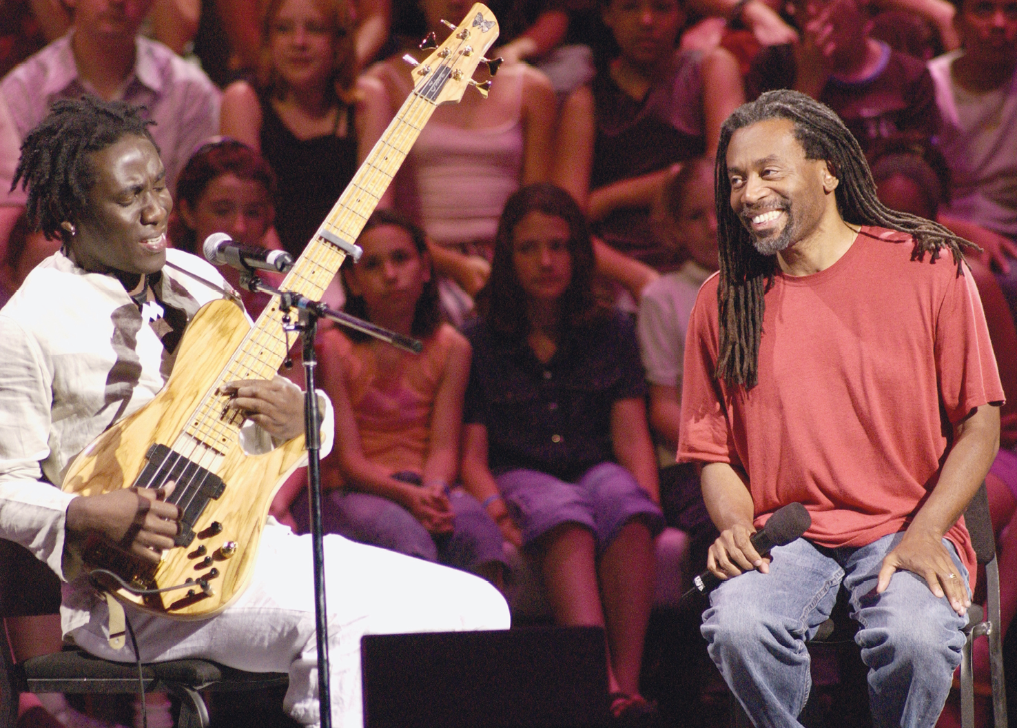 Fotografia. À esquerda, Richard Bona, um homem negro com camisa e calças brancas, sentado em um banco,  segurando uma guitarra. Diante dele, um microfone. Ao seu lado, Bobbu McFerrim, um homem negro, cabelos compridos com tranças, usando uma camiseta vermelha e calça jeans. Ele está sentado, segurando um microfone com a mão direita e sorri. Ao fundo, plateia com algumas pessoas sentadas.