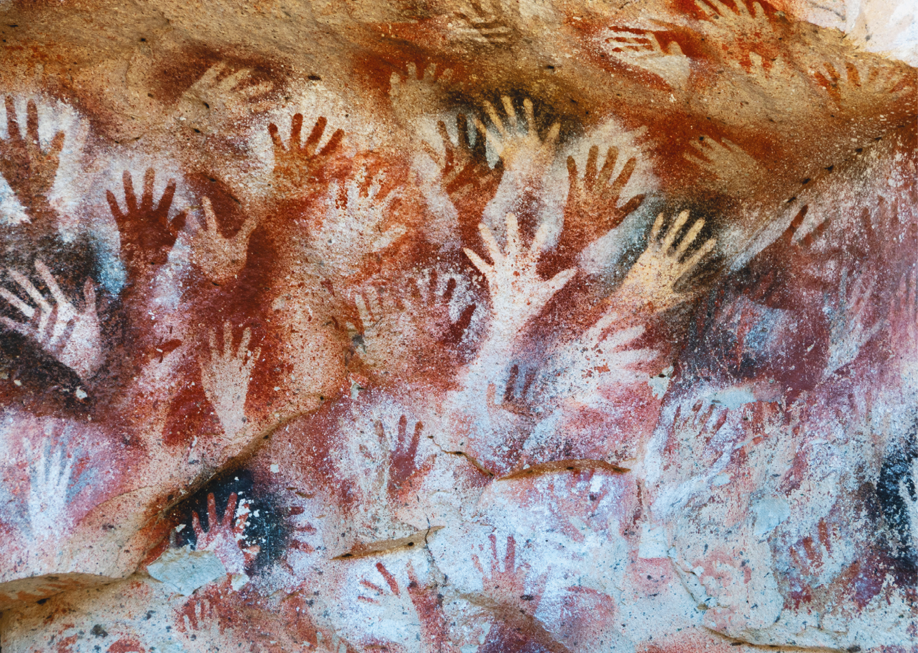 Fotografia. Pintura rupestre. Diversas mãos, algumas claras e outras escuras, impressas em uma rocha.