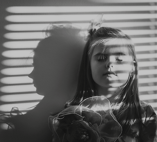 Fotografia em preto e branco. Uma menina com os olhos fechados; sobre o seu rosto, faixas horizontais da sombra de uma persiana. Atrás da menina, na parede, a sombra de seu perfil.