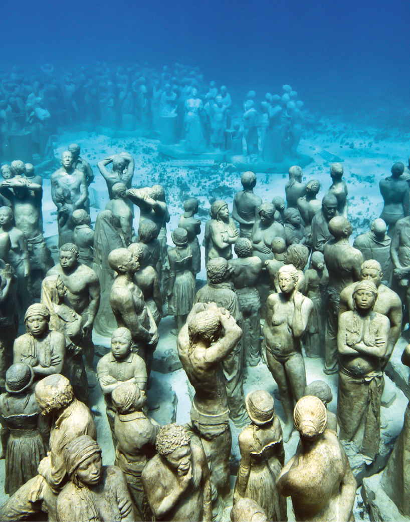 Fotografia. Diversas esculturas de pessoas dispostas em um leito arenoso no fundo do mar, de águas azuladas e transparentes,  compondo uma multidão.