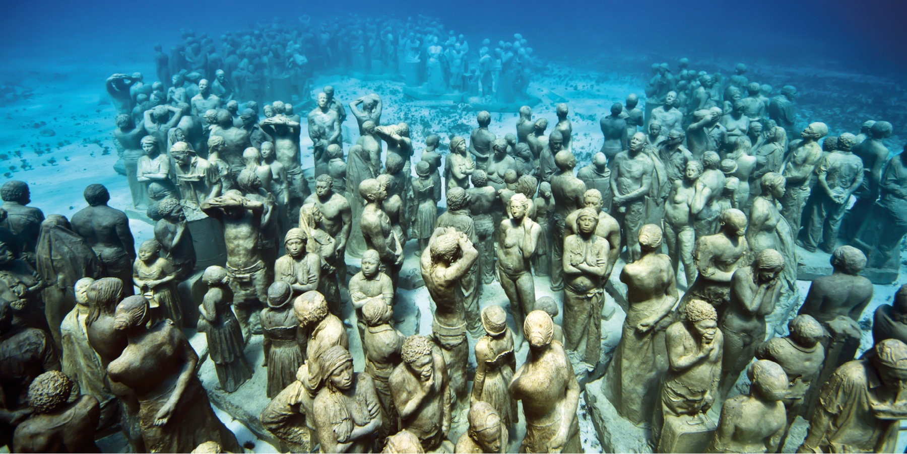 Fotografia. Diversas esculturas de pessoas dispostas em um leito arenoso no fundo do mar, de águas azuladas e transparentes, compondo uma multidão.