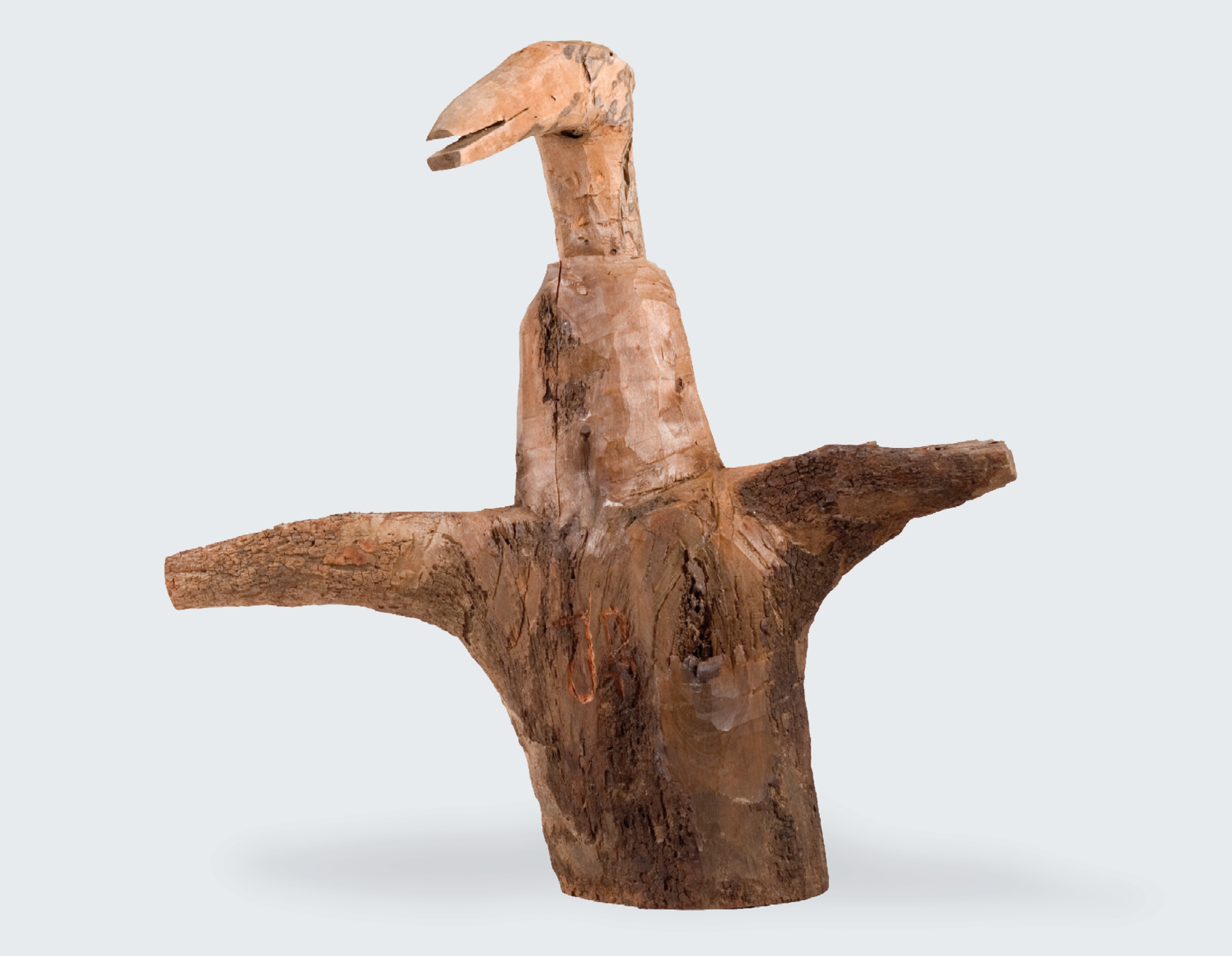Fotografia. Pedaço de um tronco de árvore com galhos esculpido em forma de uma ave de asas curtas abertas e pescoço e bico longos.