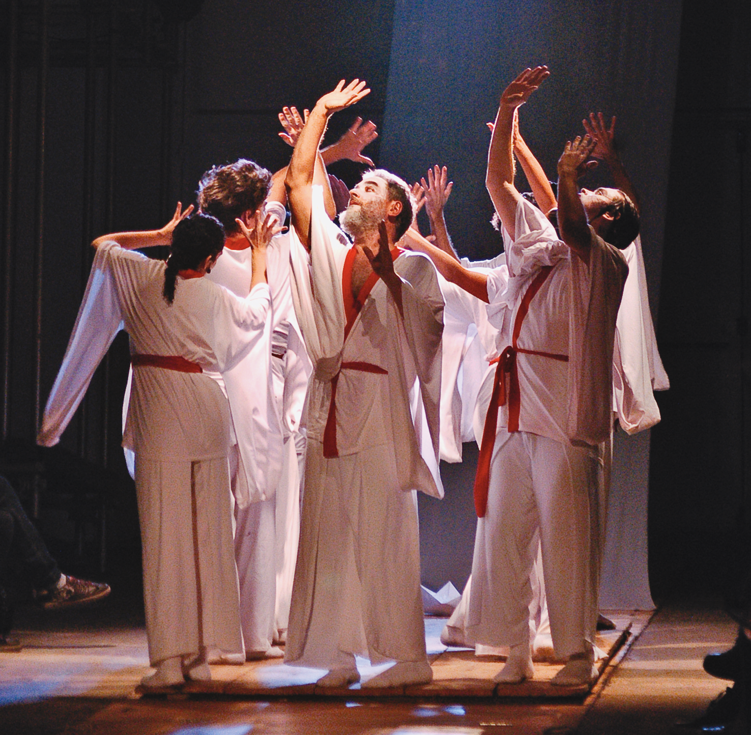 Fotografia. Espetáculo de teatro. Um grupo de atores vestindo quimono branco com faixas vermelhas em torno da cintura. Eles estão em pé sobre um tablado de madeira, com os braços para cima e as mãos espalmadas. A cena é iluminada por um facho de luz.