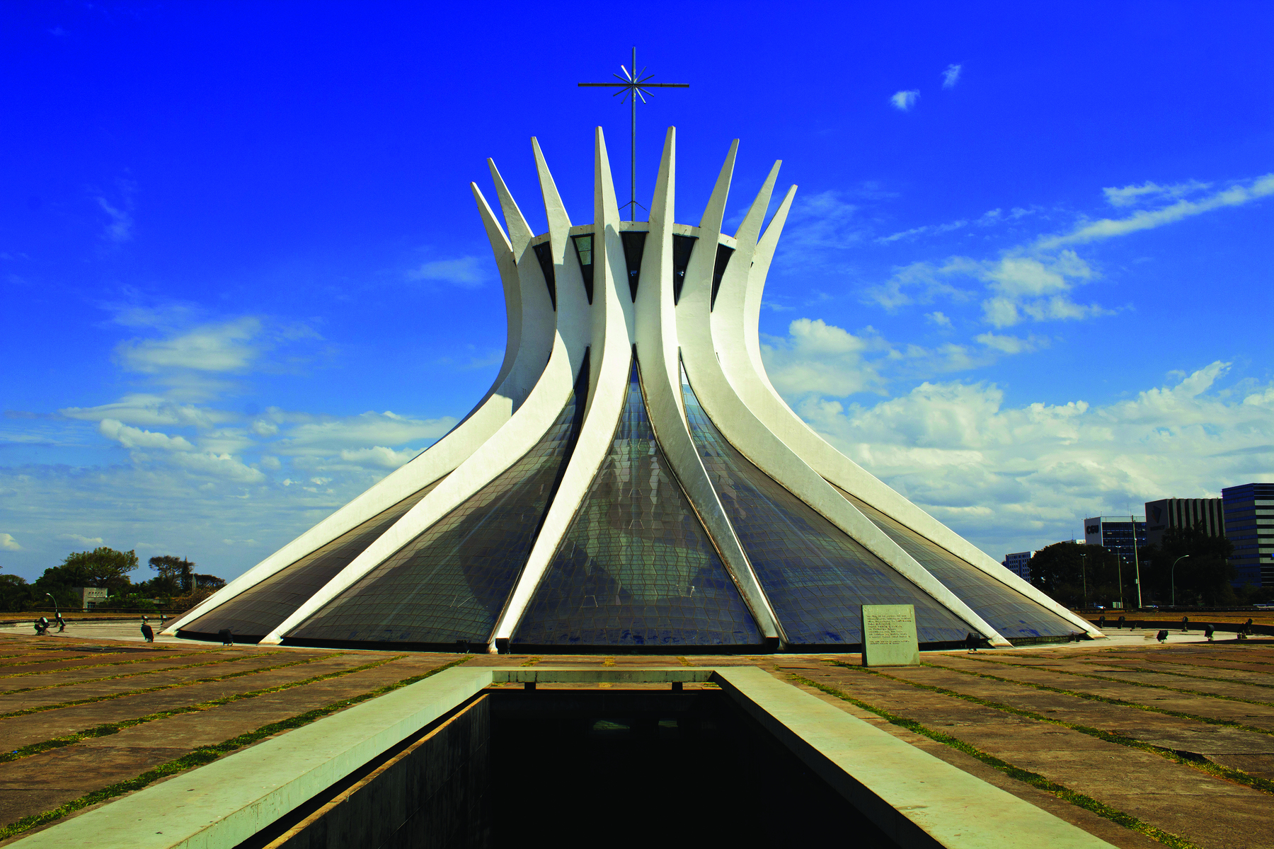 Fotografia. Construção em formato de cúpula hiperbólica, com colunas brancas, de extremidades afiladas, recurvadas e que se tocam. No topo há uma cobertura em forma circular encimada por uma estrutura metálica em formato de estrela. À frente, em uma esplanada cinza entremeada por vegetação, há um espelho de água retangular circundado por uma faixa branca. Ao fundo há árvores e outros edifícios, sob um céu azul com nuvens brancas.