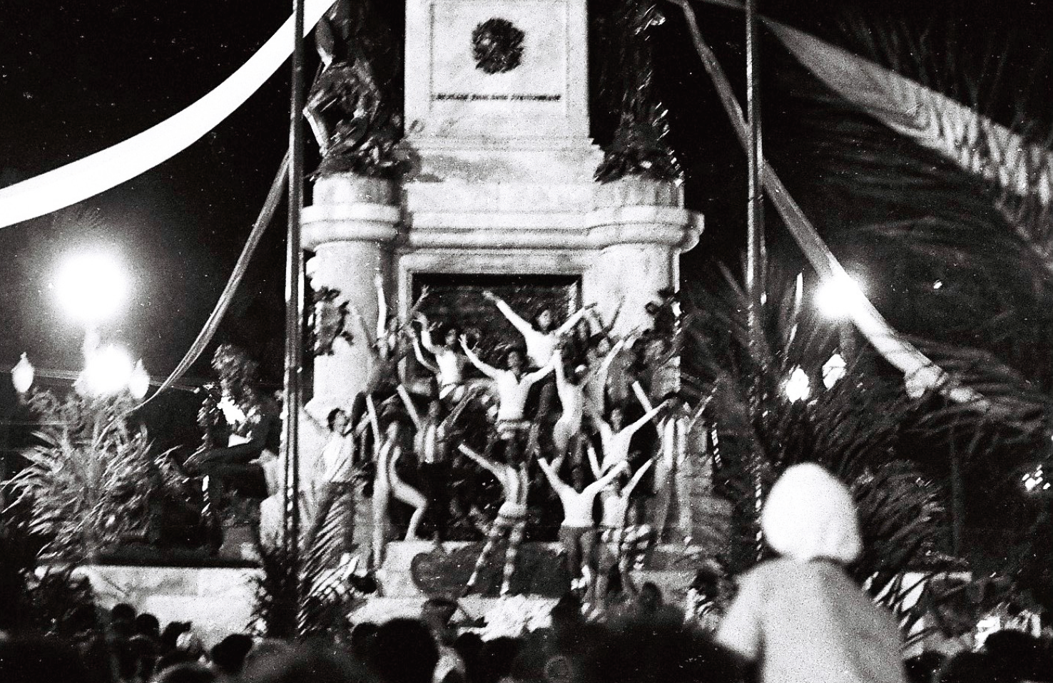 Fotografia em preto e branco. Um grupo de bailarinos com os braços estendidos para cima em uma pose. Ao fundo, um monumento. Ao redor, as pessoas de uma plateia. A cena é noturna, iluminada por algumas lâmpadas da via pública.
