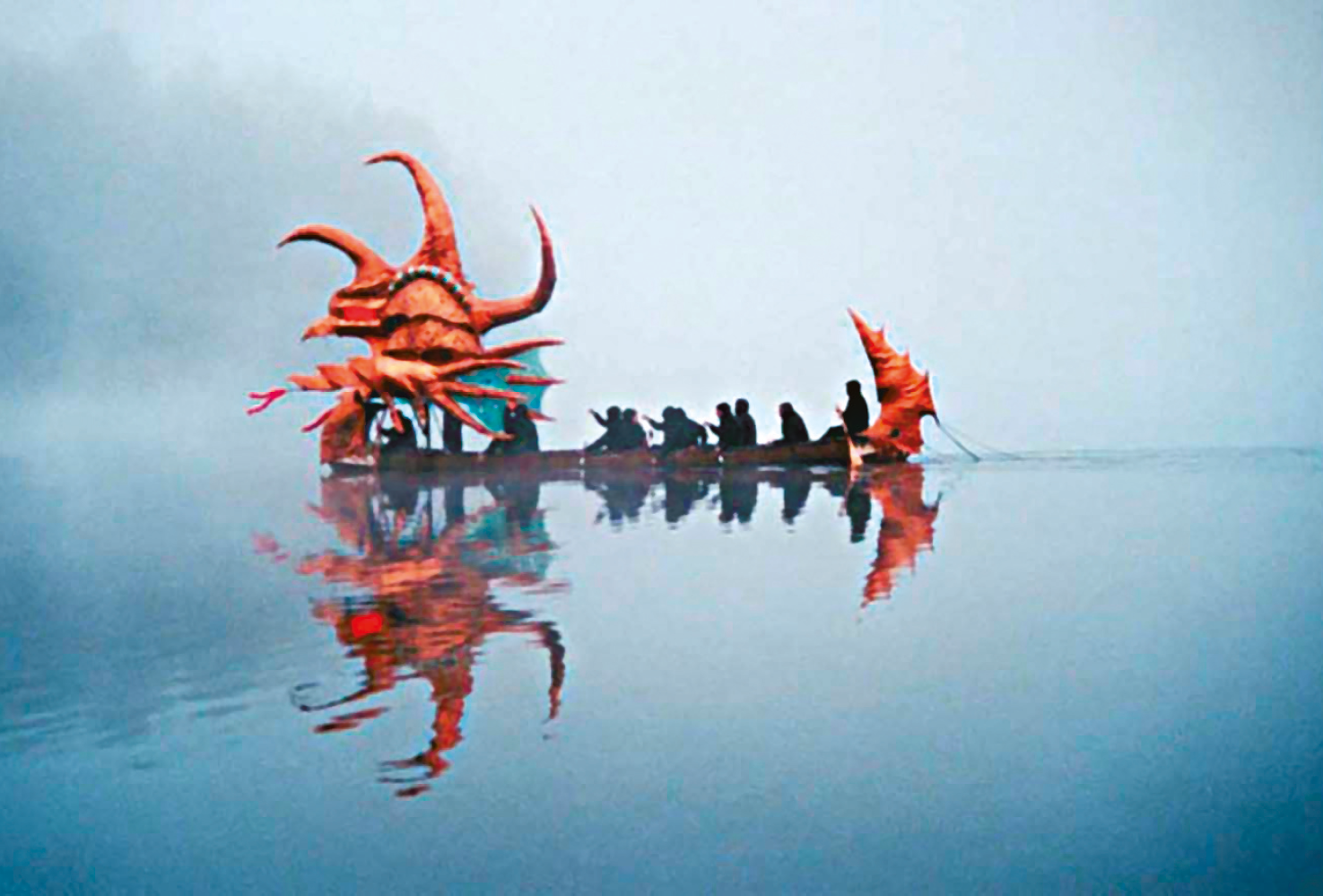 Fotografia. Águas tranquilas de um lago, com leves ondulações e reflexos da cena que se desenrola na superfície. No centro da imagem há um barco de perfil, com a proa voltada para a esquerda. Dentro dele há várias pessoas sentadas em fila, e sobre elas, na proa, há uma grande figura cenográfica na forma de um crustáceo avermelhado com três grandes chifres. Na popa do barco há uma figura cenográfica representando a cauda pontiaguda do crustáceo. Ao fundo há uma névoa espessa, através da qual aparece uma faixa de floresta densa às margens do lago.