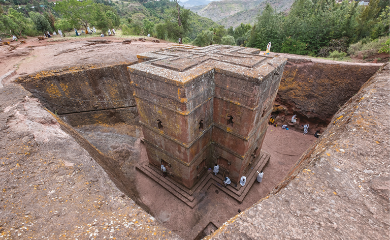 Fotografia. Construção em forma retangular composta de dois prédios conectados formando uma cruz, no interior de uma abertura escavada na pedra. Dentro da abertura, na base da construção, há algumas pessoas. Ao fundo, vegetação e algumas montanhas.