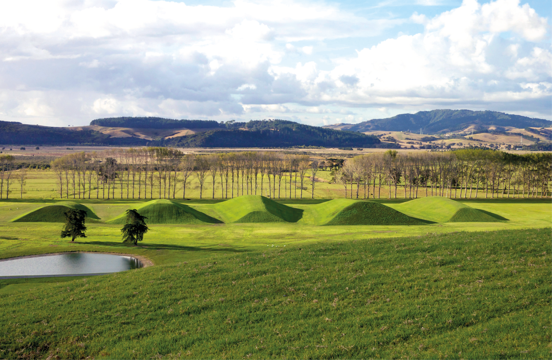 Fotografia. Imagem de uma obra de arte feita com elementos naturais em meio à paisagem natural. À esquerda há um pequeno lago em um terreno totalmente coberto de grama. Atrás dele há duas árvores pequenas lado a lado, em frente a uma construção ondulada coberta de grama. Atrás da ondulação, diversas árvores com copas rasas alinhadas. Ao fundo, montanhas com vegetação, e acima, céu com muitas nuvens brancas espessas e algumas faixas azuis.