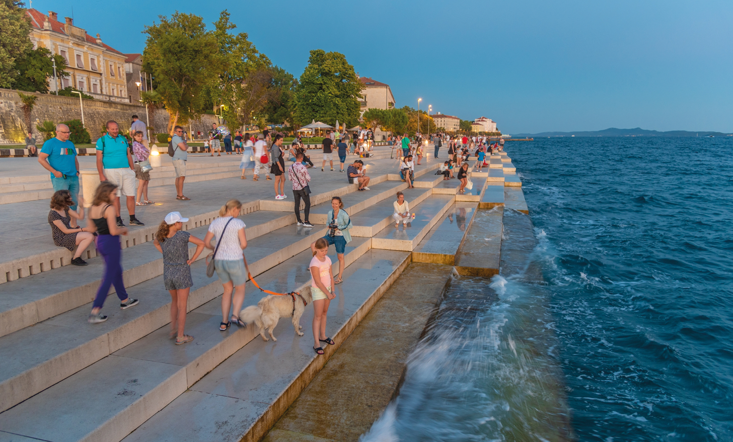 Fotografia. Paisagem com parte de uma cidade à beira do mar. À esquerda,  há uma faixa larga e extensa revestida de calçamento claro com diversos degraus, construídos à beira do mar. Sobre o calçadão,  um edifício grande e outros menores. Ao lado do edifício maior, árvores. Sobre os degraus,  há dezenas de pessoas, algumas em pé e outras sentadas. As ondas quebram em alguns desses degraus. Do lado direito, o mar. Ao fundo, há uma faixa distante de terra firme e um céu azul e sem nuvens.